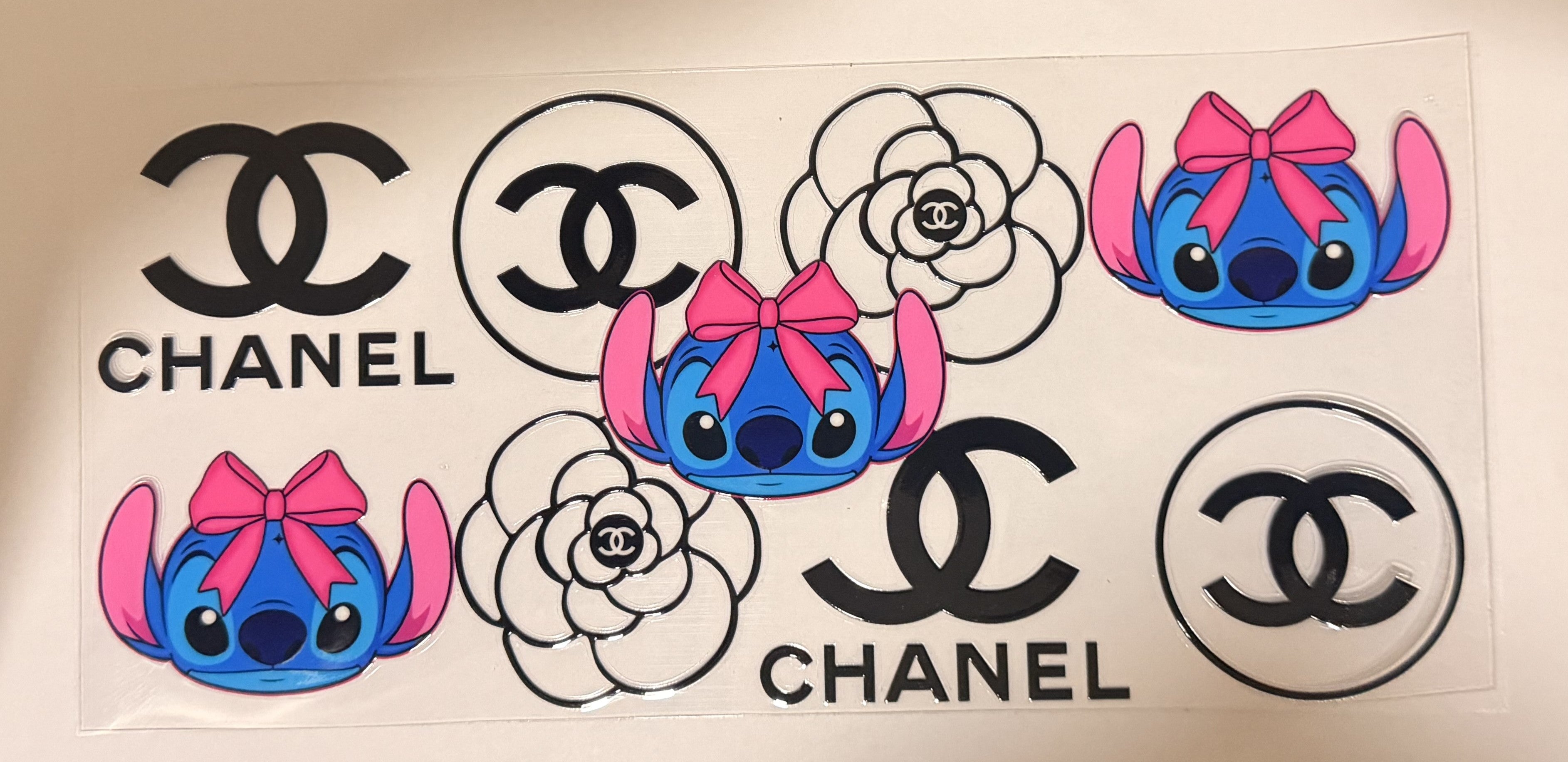 A sheet of temporary decals featuring Chanel logos and Stitch characters. The decals include black and white Chanel logo designs, white camellia flowers with Chanel CC inside, and blue Stitch characters wearing pink bows.