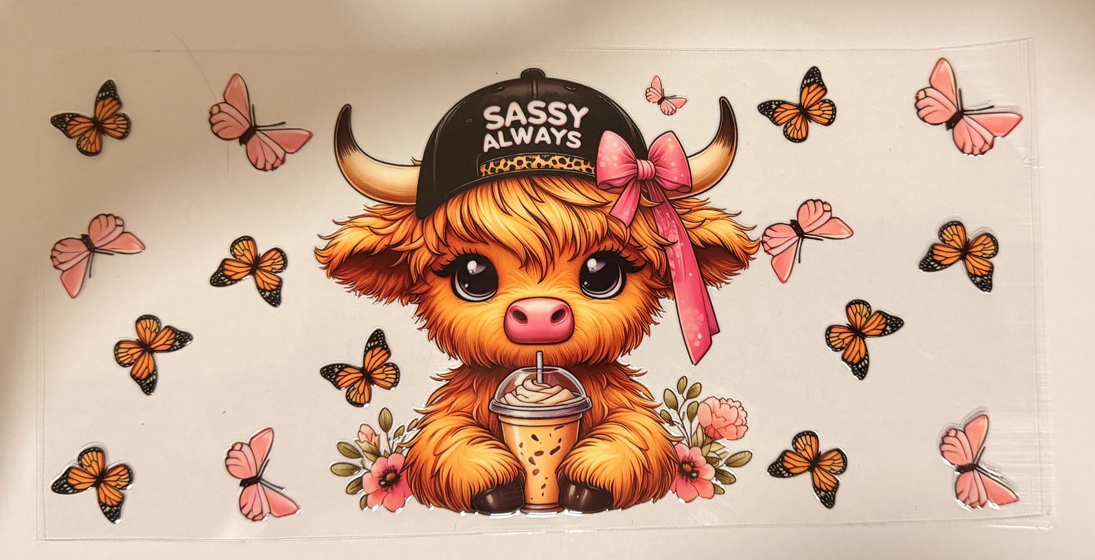 A cartoon highland cow wearing a black cap that says 'Sassy Always' and a pink bow, holding a drink, surrounded by pink and orange butterflies on a white background.